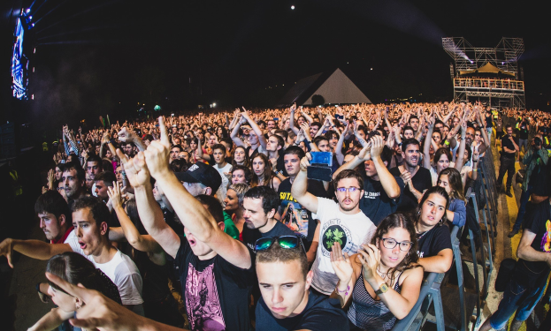 Menschenmenge auf einem Musikfestival mit erhobenen Händen, einige sitzen auf Stühlen und andere stehen, begrenzt von einem Metallzaun, mit Gebäuden und Lichtern im Hintergrund unter einem dunklen Himmel.