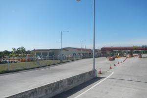 Straße mit Verkehrskegeln, Metallzaun, Gruppe von Gebäuden mit Fenstern, Straßnpöle, Hinweisschild, Brücke, Fahrzeuge, Bäume und bewölkter Himmel.