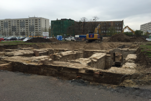 Großer Erdberg auf einer Baustelle umgeben von Baggern, Fahrzeugen, Gras, Bäumen und Gebäuden mit Fenstern in einem bewölkten Himmel über einer Straße.