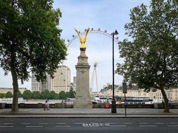 Das London Eye im Hintergrund mit einer Statue auf einem Sockel im Vordergrund, eine Straße, Bäume, ein Lichtmast, Menschen auf einem Fußweg, Schiffe auf dem Wasser und Gebäude unter dem Himmel.