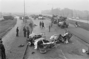 Schwarz-weiß-Szene eines Autounfalls am Straßenrand mit mehreren Fahrzeugen, einer Gruppe von Menschen in der Nähe und Hintergrundelementen wie Laternenpfähle, Bäume und Himmel.