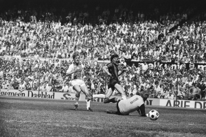 Schwarzes und weißes Foto von Fussballspielern auf einem Feld während eines Bundesligaspiels zwischen Ajax und Hannover 96 in der Saison 1978-79, mit Zuschauern und einem Banner im Hintergrund.