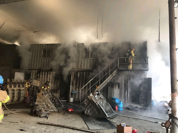Feuerwehrleute in Helmen verwenden Schläuche, um ein Lagerfeuer zu löschen, mit Rauch, der aus dem Gebäude aufsteigt, Treppen mit Geländern, die zum Eingang führen, einem angeketteten Pfahl auf der rechten Seite, Fenstern an einer Wand im Hintergrund und verstreuten Gegenständen wie Kartons und Rohren auf dem Boden.