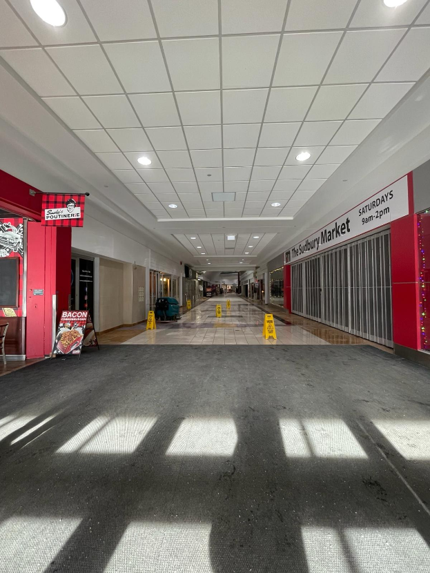 Innenseite eines Einkaufszentrums mit Deckenbeleuchtung, einem Stuhl und einer Tafel mit der Aufschrift "Sydbury Market - Vorher und Nachher" links und verstreuten Gegenständen rechts und auf dem Boden.