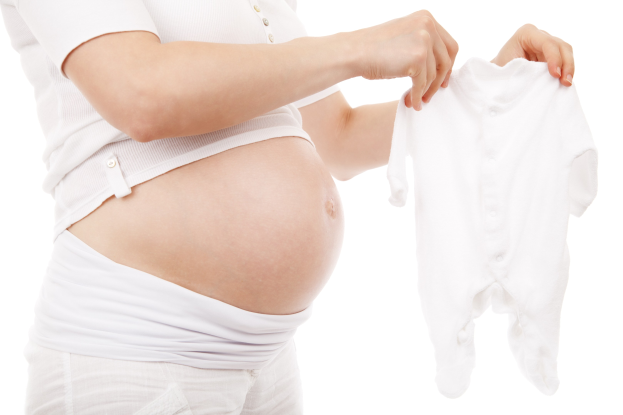 Schwangere Frau in einem weißen Kleid, die ein weißes Babyzäpfchen hält, mit einem zufriedenen Gesichtsausdruck.
