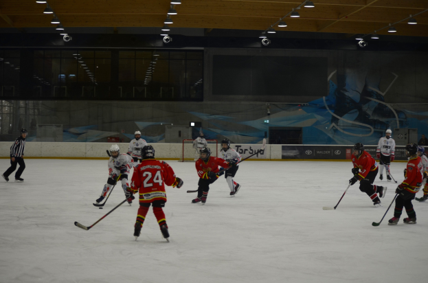 Gruppe von Menschen in Helmen und Eishockey-Ausrüstung, die auf einer Indoor-Eisfläche mit einer bemalten Wand und heller Deckenbeleuchtung Eishockey spielen.