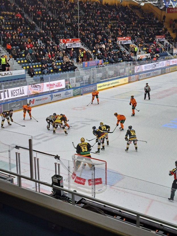 Eine Gruppe von Menschen spielt Hockey auf einem Eisstadion, mit einem Netz, einer Umzäunung, sitzenden Zuschauern, Bannern und Scheinwerfern von oben.