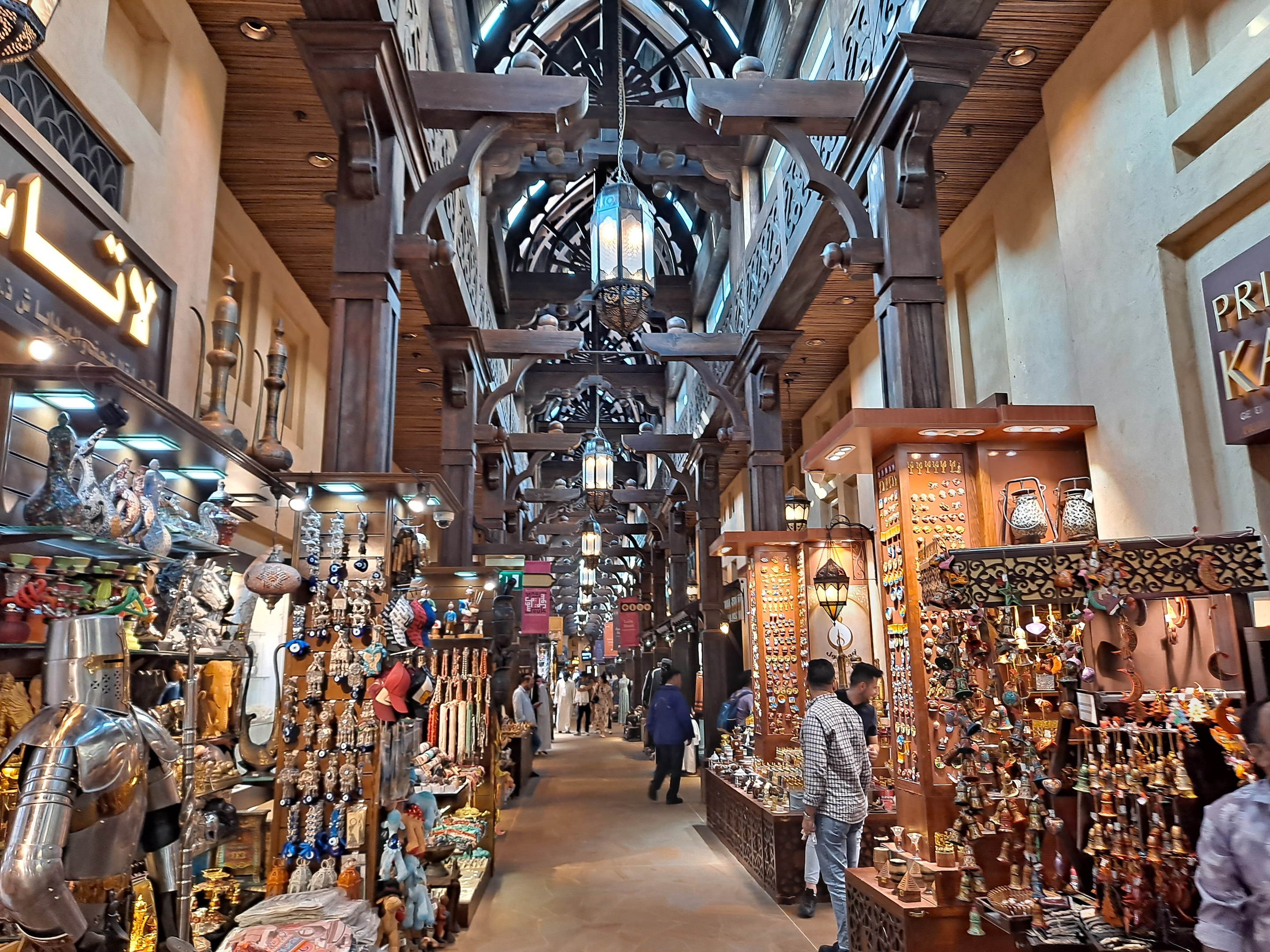 Ein geschäftiges Dubai-Bazar mit Menschen, die zwischen Regalen voller Artikel hindurchgehen, Wänden mit Texttafeln und einer Decke mit warmem Licht.