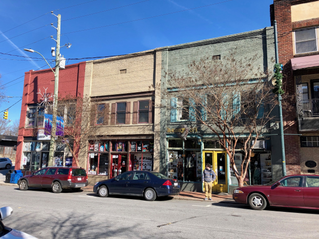 Eine Stra├če in der Stadt mit geparkten Autos, Geb├Ąuden, B├Ąumen, Laternen, Strommasten mit Kabeln und einer Person, die auf dem Gehweg geht, unter einem klaren Himmel, darstellend eine gewerbliche Immobilie zum Verkauf f├╝r $1.250.000.