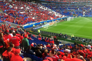 Ein volles Stadion mit Sitz- und Stehplätzen, das ein Fußballspiel verfolgt, mit Bannern, einem Torpfosten und Deckenleuchten.