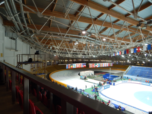 Große Indoor-Eisbahn mit zahlreichen Menschen, die Schlittschuh laufen, umgeben von Sitzplätzen, Geländern, Brettern, Beleuchtungskörpern und strukturellen Elementen, mit einer Wand im Hintergrund.