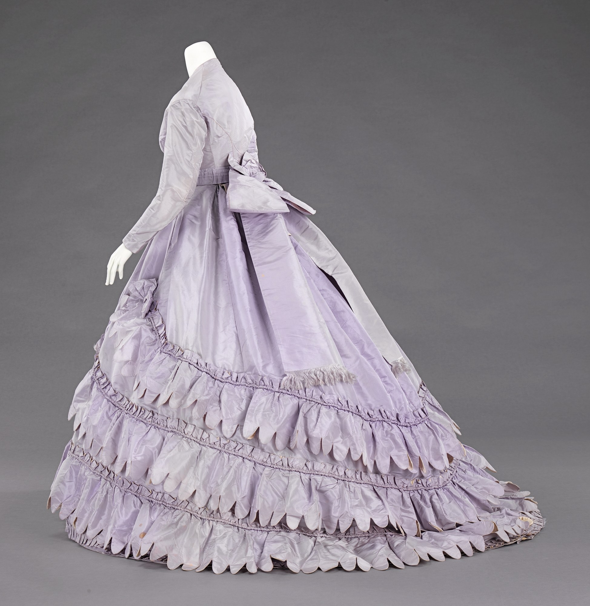 Eine Schaufensterpuppe in einem lila Ballkleid mit engem Oberteil, vollem Rock und langen Ärmeln vor einem grauen Hintergrund.
