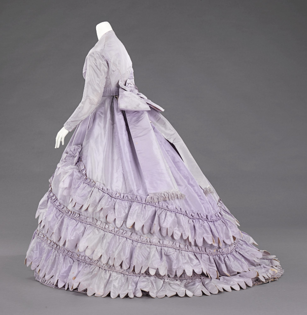 Eine Schaufensterpuppe in einem lila Ballkleid mit engem Oberteil, vollem Rock und langen Ärmeln vor einem grauen Hintergrund.