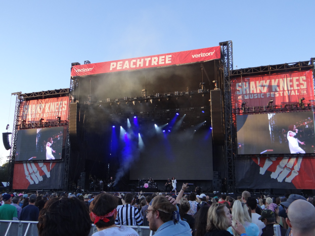 Eine große Menschenmenge steht vor einer hell erleuchteten Bühne beim Shaky Knees Music Festival, auf der Musiker unter Bannern mit Text spielen, mit einem Geländer im Vordergrund und einem klaren blauen Himmel im Hintergrund.