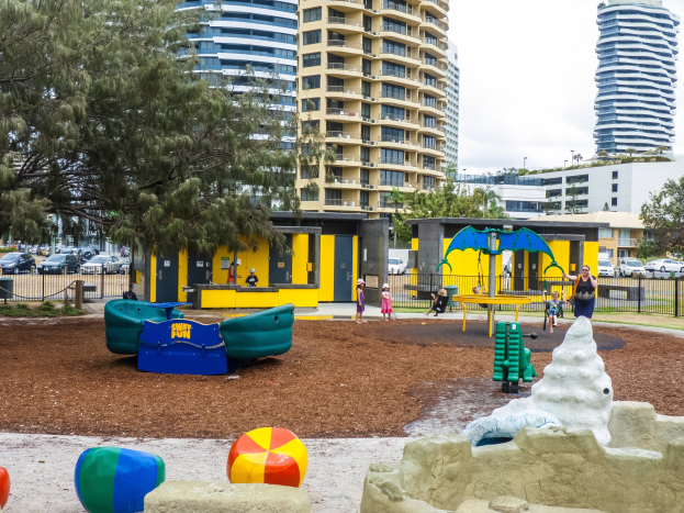 Spielplatz mit Kindern auf Geräten, umgeben von Bäumen und Geländern, in einer städtischen Umgebung mit hohen Gebäuden, Fahrzeugen und sichtbarem Himmel.