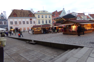 Ein geschäftiger Weihnachtsmarkt in Tallinn, Estland mit Menschen um dekorierte Stände, festliche Lichter, Gebäude im Hintergrund, bewölkter Himmel, Treppe zum Markt und Töpfe mit Pflanzen herum.