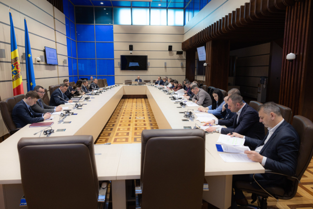 Eine Gruppe von Menschen um einen langen Tisch versammelt mit Papieren, Büchern, Mikrofonen und anderen Gegenständen während einer Sitzung des Europäischen Parlaments in Brüssel, mit Flaggen und Bildschirmen im Hintergrund.