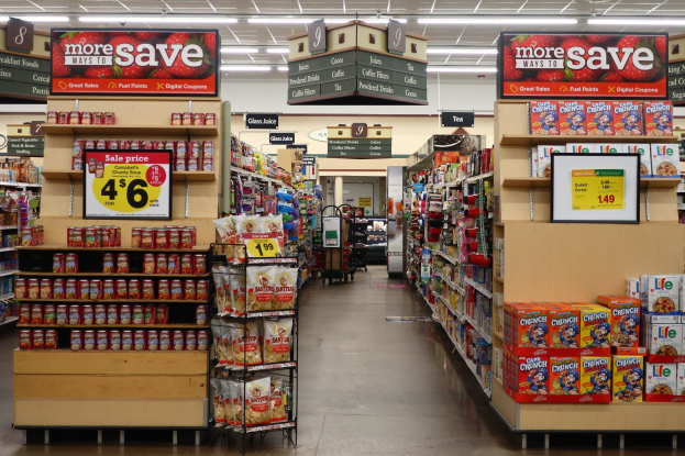 Ein Kiosk-Innenraum Mit Verschiedenen Lebensmitteln, Die Auf Regalen, Regalen Und Dem Boden Angezeigt Werden, Einschl. Beleuchteter Deckenlichter Und Informationsschilder Im Hintergrund.