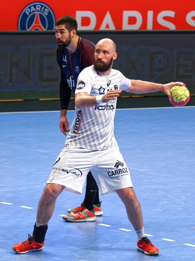 Zwei Männer spielen Handball auf einem Court, einer hält einen Ball, mit einem Hintergrundboard, das 'Paris Saint-Germain vs Paris Saint-Germain' anzeigt.