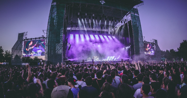 Eine gro├če Menschenmenge auf einem Musikfestival schaut auf einer beleuchteten Bühne mit Instrumenten und Bildschirmen, die vor einem Geb├Ąude, B├Ąumen und einem klaren blauen Himmel steht.