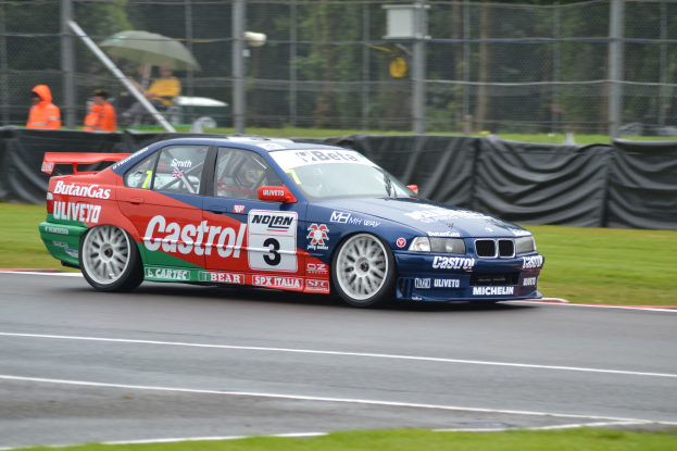 Ein Auto fährt auf einer Rennstrecke mit Gras auf beiden Seiten, mit einem Maschendrahtzaun, der mit schwarzem Stoff bedeckt ist, und Menschen in orangefarbenen Jacken mit Schirmen im Hintergrund.