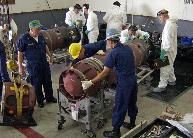Eine Gruppe von Männern in Helmen und Handschuhen bei der Entschärfung einer großen Bombe in einer Fabrik mit Maschinen und einer textbedeckten Wand im Hintergrund.