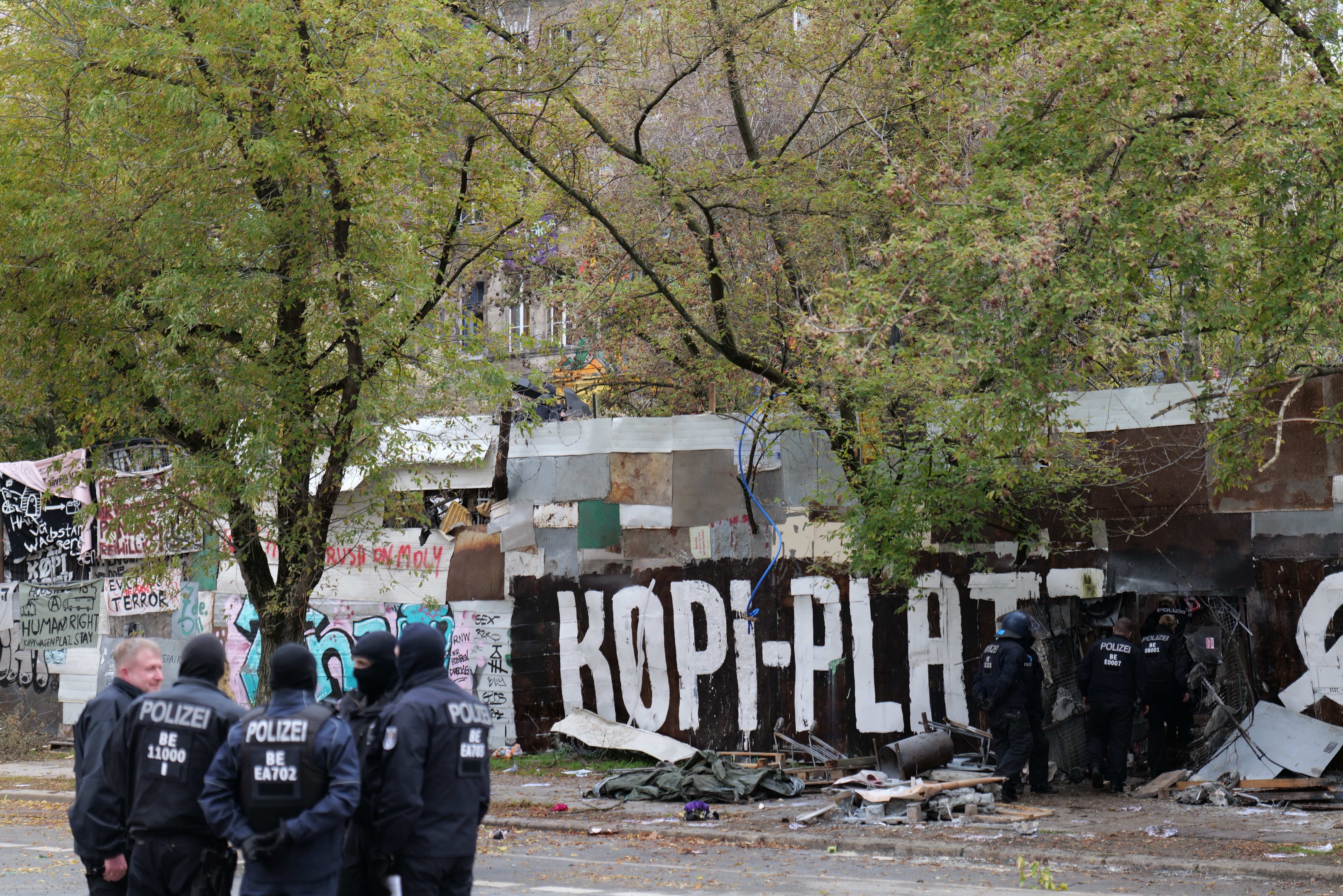 Polizeibeamte in schwarzen Uniformen und Helmen stehen vor einem mit Graffiti bedeckten, baufälligen Gebäude mit verstreuten Trümmern und Bäumen im Hintergrund.