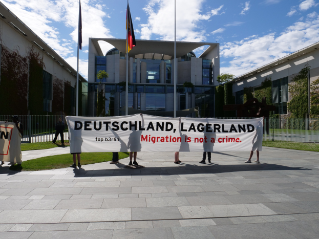 Gruppe von Menschen mit einem Transparent "Deutschland, Lagerland Migration ist kein Verbrechen" vor einem Gebäude mit Fenstern, umgeben von Gras, einem Metallzaun, Pflanzen, Bäumen und Fahnenmasten bei einem bewölkten Himmel.
