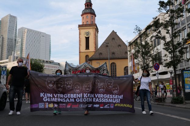 Eine Gruppe von Menschen in Masken, die eine Plakette tragen und die Straße entlanggehen, mit einem geparkten Auto auf der linken Seite, Gebäuden und einem Kirchturm im Hintergrund unter einem klaren blauen Himmel.