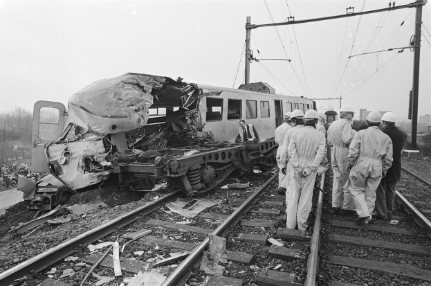 Ein Schwarz-Weiß-Bild von einer Zugkollision auf den Schienen mit Menschen in Helmen drumherum, vor Elektromasten, Bäumen, Gebäuden und einem klaren Himmel.