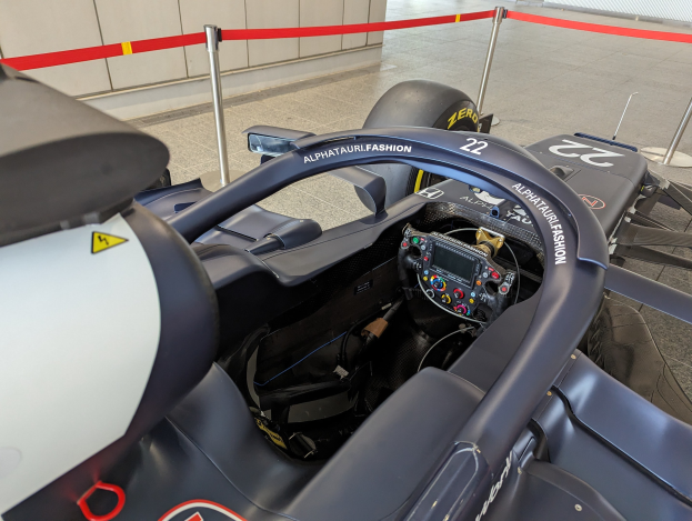 Cockpit eines Formel-E-Autos in einer Ausstellung mit einem Lenkrad im Vordergrund, einer Wand im Hintergrund und um das Auto herum abgesteckte Pfosten.