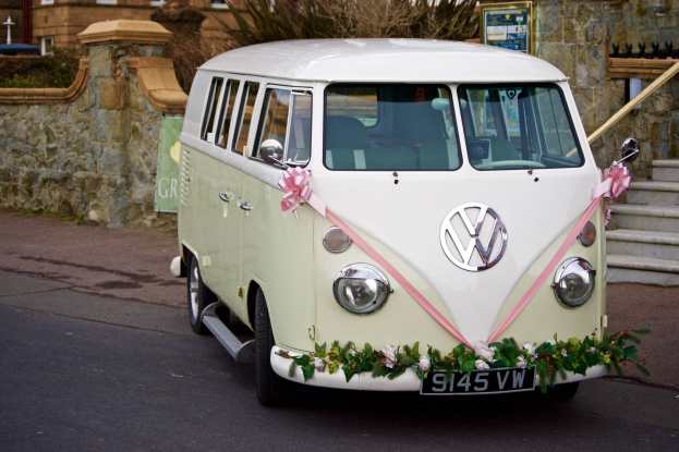 Weißer und pinker VW Camper mit floralen Verzierungen vor einer Steinmauer geparkt, neben Treppen und einem Gebäude.