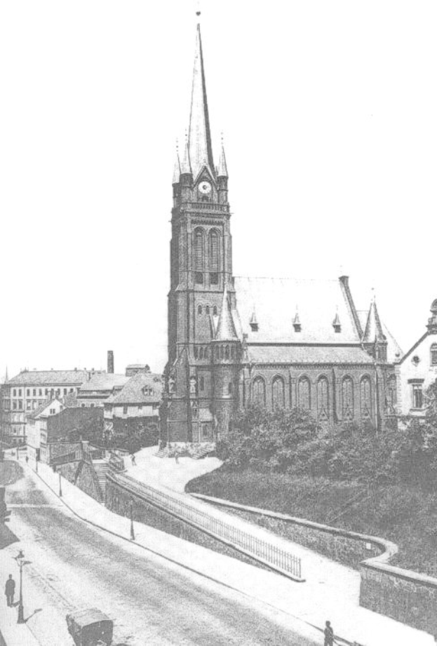 Schwarzes und weißes Foto einer Stadtstraße mit der St. Peter Kirche in Wiesbaden, Deutschland im Hintergrund, gesäumt von Gebäuden, Bäumen, Fahrzeugen und Passanten.