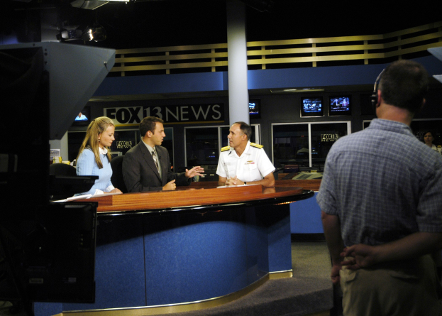 Eine Gruppe von Menschen steht um einen Schreibtisch in einem Nachrichtenstudio mit Papieren und Gegenständen darauf, einem Monitor links und Bildschirmen, einer Säule und Deckenleuchten im Hintergrund.