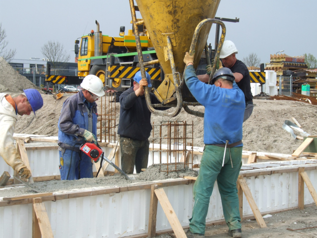Bauarbeiter mit Helmen bedienen einen Betonmischer auf einer Baustelle mit Fahrzeugen, Bäumen, Gebäuden und einem klaren Himmel im Hintergrund.