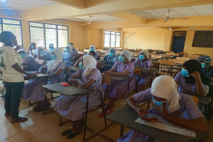 Ein Klassenzimmer in Nigeria mit Schülern, die Gesichtsmasken tragen und auf Bänken sitzen oder stehen, mit Büchern und Gegenständen auf den Tischen, Fenstern, einer Tür und Deckenlampen und Ventilatoren im Hintergrund.