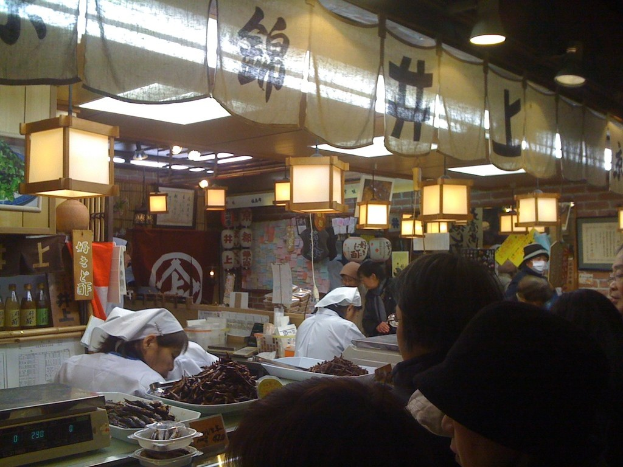 Ein Restaurantmitarbeiter und Kunden interagieren an einem Tresen mit Gegenständen, während Flaschen mit Soßen, Menüs, Kleidung und Lampen im Hintergrund zu sehen sind.