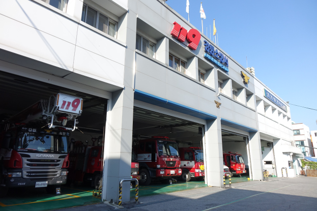 Feuerwache mit mehreren Feuerwehrautos vor der Tür, mit Fenstern, Säulen und Namensschildern, mit Fahnen im Vordergrund und anderen Gebäuden unter einem klaren blauen Himmel.