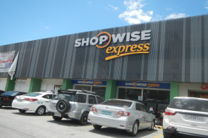 Ein Shopwise Express-Laden mit parkenden Autos davor, mit einer Banner auf der linken Seite, unter einem bewölkten Himmel.