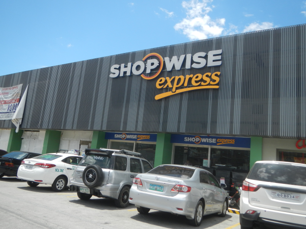 Ein Shopwise Express-Laden mit parkenden Autos davor, mit einer Banner auf der linken Seite, unter einem bewölkten Himmel.