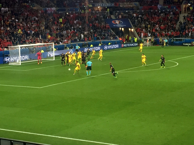 Eine Gruppe von Menschen, die auf einem Feld in einem Stadion Fußball spielen, mit einem Netz auf der linken Seite, einer Zuschauermenge auf den Rängen und beleuchteten Tafeln im Hintergrund.