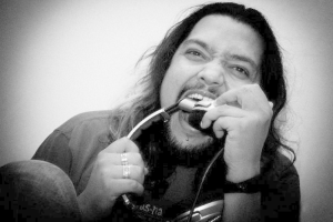 Ein Mann mit langen Haaren und Bart singt leidenschaftlich in ein Mikrofon, trägt ein schwarzes T-Shirt und Jeans vor einer Wand.