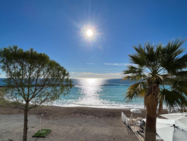 Ein sonniger Strand mit Palmen, Sonnenschirmen und üppiger Vegetation an der französischen Riviera unter einem strahlend blauen Himmel.