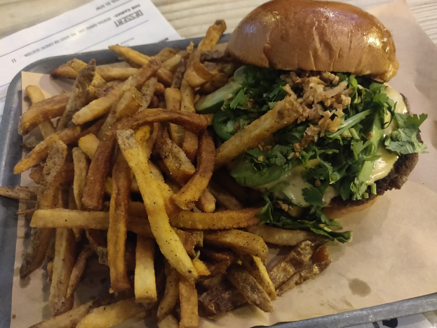 Ein Tablett mit einem Hamburger, der mit Salat, Tomate und Zwiebeln belegt ist und goldenen, knusprigen Pommes frites, das auf einem Tisch mit einem Blatt Papier daneben steht.
