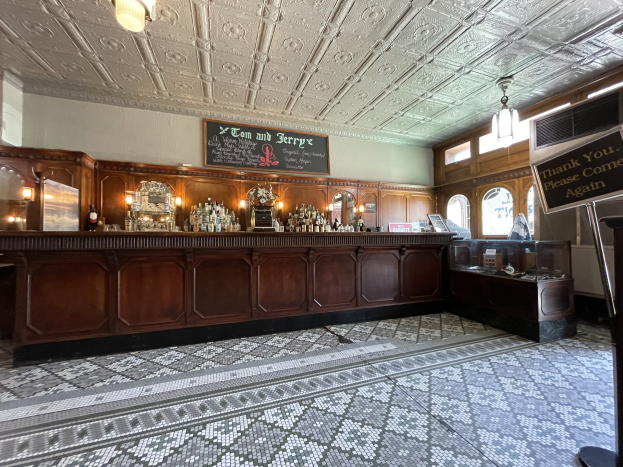 Ein großer Raum mit einer zentralen Bar, Deckenleuchten, Flaschen auf einem Tisch und einer Schiefertafel auf der rechten Seite, im Tom and Jerry Pub in London.