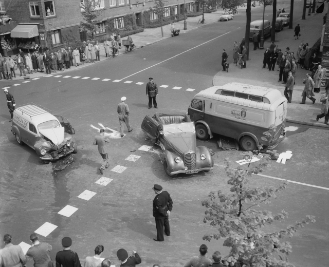 Schwarz-weißes Bild eines Autounfalls an einer Straßenkreuzung mit einem Van, anderen Fahrzeugen und einer Gruppe von Menschen in der Nähe, mit Gebäuden, Bäumen, Pfählen und Texttafeln im Hintergrund.