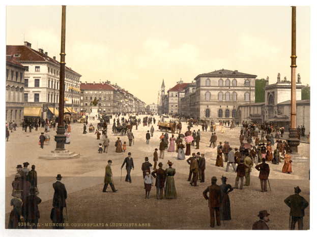 Schwarz-weiß-Foto einer belebten Münchner Straße in den frühen 1900er-Jahren mit Fußgängern, einem Pferdewagen, Gebäuden, Bäumen und einem sichtbaren Himmel.