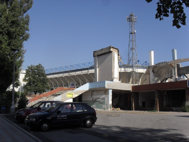 Alter Stadion mit parkenden Autos davor, umgeben von Gebäuden, Bäumen, Strommasten und einem Turm unter einem sichtbaren Himmel.