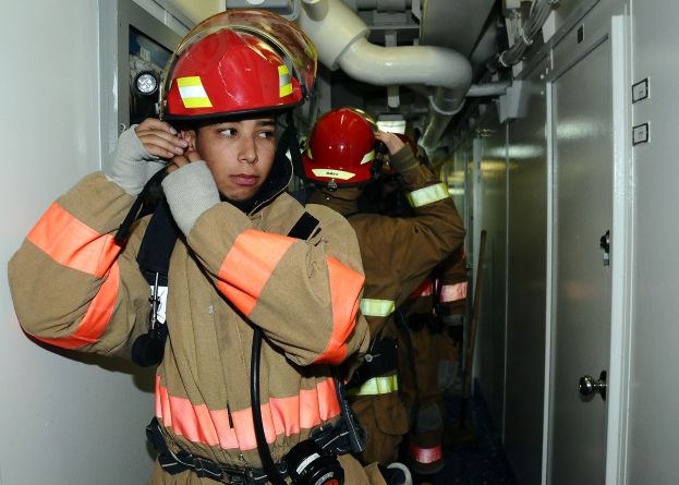 Feuerwehrleute in Uniform stehen in einem Raum mit einer Tür auf der rechten Seite und Rohren im Hintergrund während einer Übung.