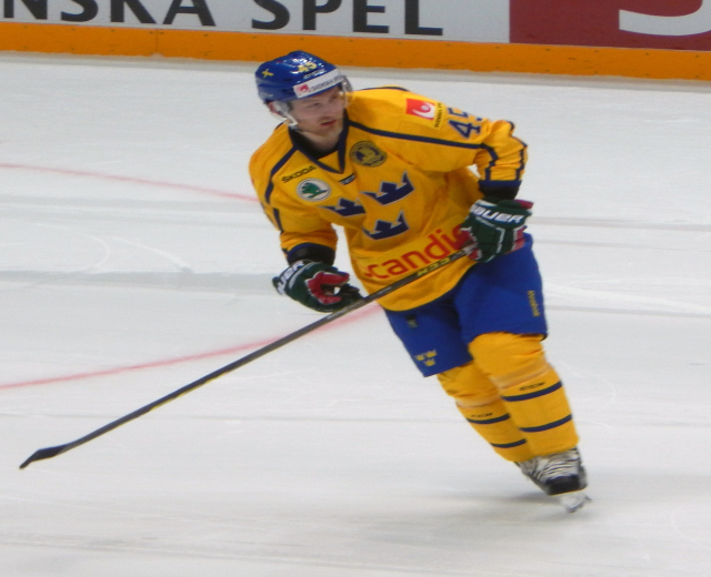 Eishockeyspieler in Bewegung auf dem Eis, trägt Helm, Handschuhe und Schlittschuhe, hält einen Hockeystock und hat ein Schild mit Text im Hintergrund.